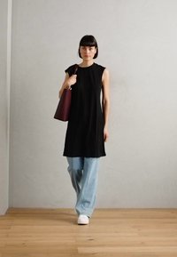 Tunica nera senza maniche, vestibilità comoda, abbinata a jeans a gamba larga blu chiaro e sneakers bianche. Tiene una borsa di un profondo borgogna. Sfondo in legno.