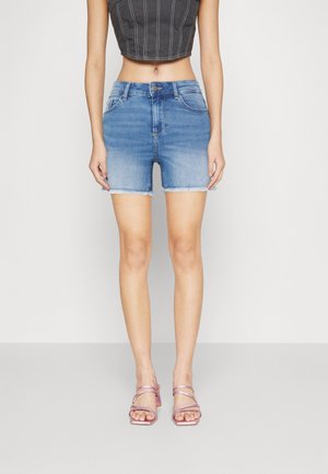Donna che indossa pantaloni corti in denim blu a vita media con orlo sfrangiato, un top nero crop e sandali con tacco rosa, in piedi su uno sfondo semplice.