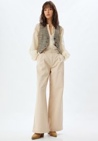 Beige wijde broekjes gecombineerd met een licht, doorschijnend blouse en een houndstooth vest. De outfit heeft subtiele texturen en neutrale tinten.