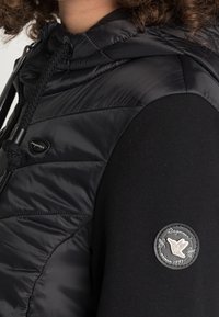 Gilet imbottito nero con finitura testurizzata e cappuccio, abbinato a una manica nera aderente con un patch logo circolare.
