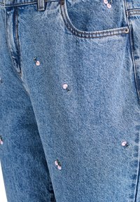 Svetlo modri traper hlače z razpršenimi roza cvetovi češnjevega cveta vezeninami. Teksturovana tkanina s klasičnim dizajnom petih žepov in kovinsko gumbom.