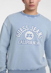 Šviesiai mėlynas džemperis su apvalia iškirpte, kuriame yra baltas grafiškas tekstas „GUESS JEANS“ ir „CALIFORNIA“ dideliu šriftu. Minkšta medžiaga, laisvas kirpimas.