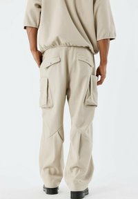 Pantalons cargo beiges à coupe ample, dotés de grandes poches latérales, d'une texture lisse et d'un léger éclat, associés à des chaussures noires.