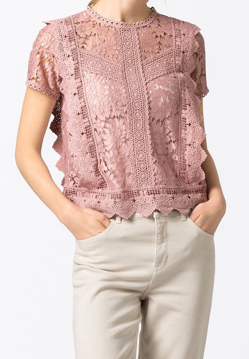 Femme portant un top en dentelle rose poudré à manches courtes avec des motifs floraux et des bords festonnés, associé à un pantalon beige clair, mains dans les poches.