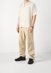 Uomo che indossa pantaloni cargo beige con polsini regolabili, una camicia leggera a maniche corte con motivo, sneakers nere e un braccialetto nero.