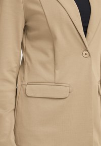 Blazer beige con un design strutturato, chiusura a un bottone, rever a punta e una tasca. Tessuto liscio con una leggera lucentezza.