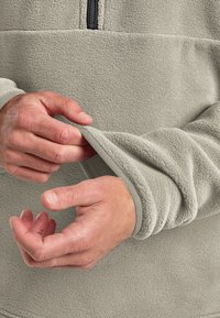 Fleece pullover in light gray with a black zipper, featuring ribbed cuffs. Hands are adjusting the sleeve, showing soft texture.