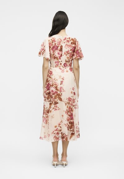 Γυναίκα με μακριά σκούρα μαλλιά, ντυμένη με ένα ανοιχτό ροζ φόρεμα με λουλούδια μέχρι τη μέση της γάμπας και μπεζ ψηλοτάκουνα, στέκεται γυρισμένη με την πλάτη προς ένα απλό φόντο.