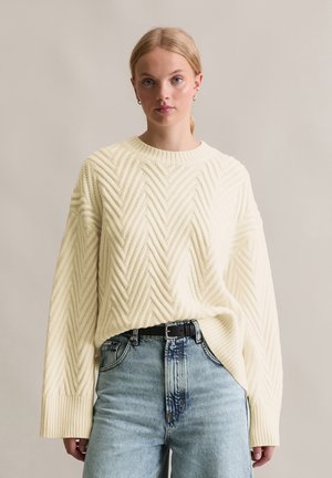 Crème kleurige sweater met een gewoven chevronpatroon, lange mouwen en een brede hals, gecombineerd met lichtblauwe high-waisted jeans en een zwarte riem.