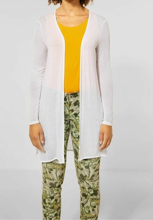 Vrouw die een lange witte cardigan draagt, een felgele top en groene broek met patroon, staand tegen een effen lichte achtergrond.