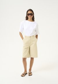 Witte T-shirt met korte mouwen, beige shorts en zwarte flip-flops. Model draagt grote zonnebril en staat met handen in de zakken tegen een neutrale achtergrond.