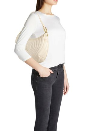 Borsa a tracolla in pelle trapuntata beige con design curvo, dettagli in oro e tasca laterale, indossata con top bianco e jeans scuri.