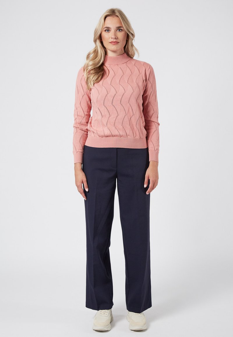Różowy sweter wykonany na drutach z wzorem falistym, z wysokim dekoltem i ściągaczami, połączony z granatowymi spodniami i białymi trampkami.