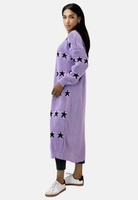 Vestido de cárdigan de punto lavanda con patrones de estrellas negras, mangas largas, puños acanalados y corte recto, combinado con zapatillas negras y blancas.