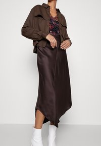 Chemisier court marron avec deux poches poitrine, superposé à un haut fleuri, associé à une jupe satinée marron avec un ourlet asymétrique et des bottes blanches.