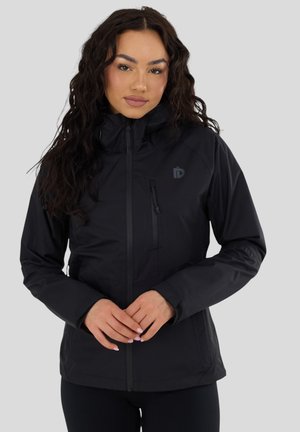 Frau mit langen dunklen lockigen Haaren, die eine schwarze Jacke mit Reißverschluss und hohem Kragen trägt, steht vor einem schlichten hellgrauen Hintergrund.