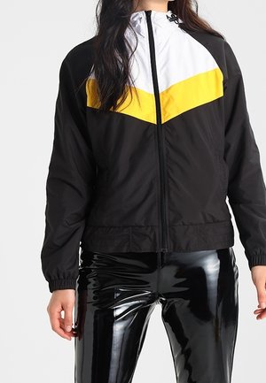 Woman wearing a black zip-up jacket with white and yellow chevron pattern, paired with shiny black vinyl pants.
