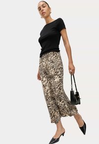 Schwarzes Kurzarmoberteil kombiniert mit beigen weit geschnittenen Hosen mit Leopardenmuster. Die Accessoires umfassen eine schwarze Handtasche und spitze Mules.