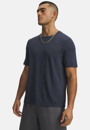 Man draagt een donkerblauw gestructureerd shirt met korte mouwen en donkergrijze korte broek, staat met ontspannen armen en kijkt opzij.