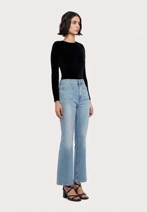 Svart fløyelslangermet topp sammen med lyseblå flagrende jeans med to frontlommer, teksturert stoff og svarte hælsandaler.