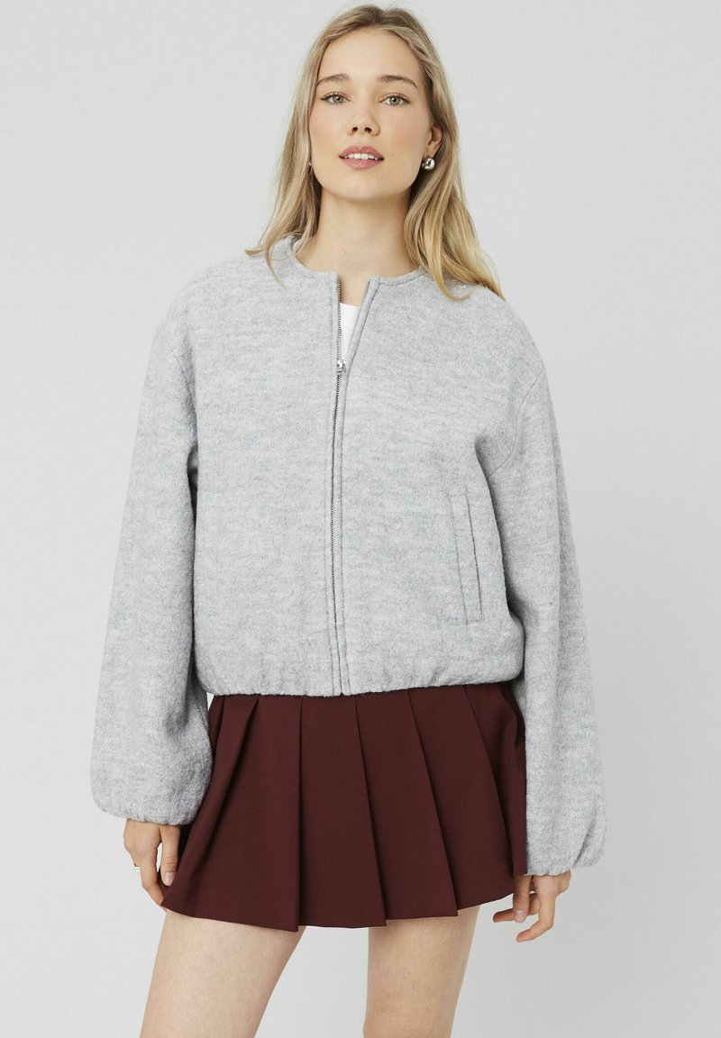 Veste bomber grise en tissu doux, avec fermeture éclair à l'avant, manches larges et poches latérales. Associée à une jupe plissée prune.