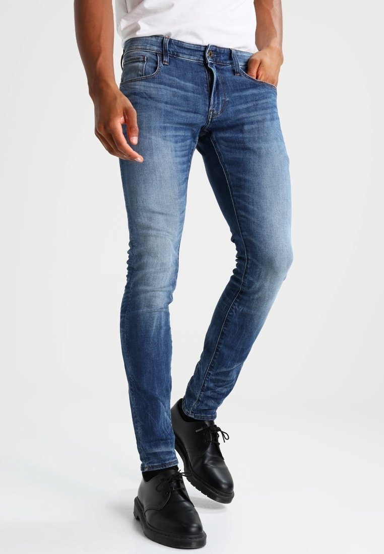 Jeans de denim azul ajustados con un aspecto desgastado y descolorido. Presenta cinco bolsillos, pasadores para cinturón y una cremallera. Combinados con zapatos negros.