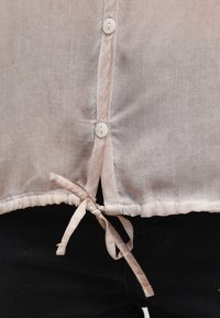 Chemise beige clair boutonnée avec une texture légèrement transparente et un ourlet froncé avec un cordon de serrage noué à la taille.