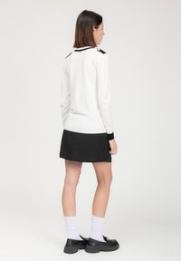 Witte trui met zwarte accenten en knopen op de schouders, gecombineerd met een zwarte rok. Model draagt zwarte schoenen en witte sokken.