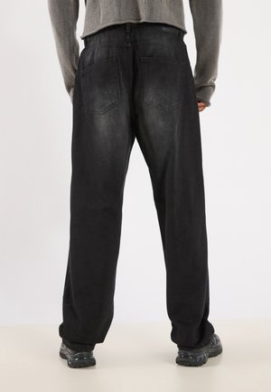 Personne portant un pantalon noir ample et des baskets foncées, vue de dos, debout sur un sol clair devant un fond uni.
