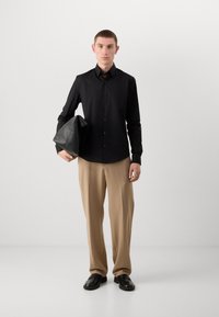 Zwart overhemd met lange mouwen, gecombineerd met beige wijde broek en zwarte schoenen, met een zwarte leren tas in de hand.