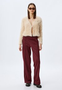 Cremefarbene Jacke aus Kunstfell mit einem cropped Design, kombiniert mit bordeauxroten, hoch geschnittenen Hosen mit glatter Textur und weitem Bein. Schwarze Schuhe.
