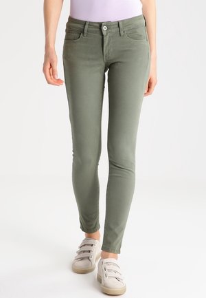 Elastyczne zielone spodnie w kolorze oliwkowym noszone z białymi trampkami na rzepy i jasnofioletowym wciągniętym topem na białym tle.