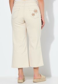 Pantalones cortos beige de corte relajado, con dos bolsillos traseros decorados con bordados de flores en naranja y blanco. Tela suave y lisa.