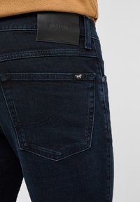 Dunkelblaue Jeans aus Denim mit glatter Textur. Merkmale sind ein Leder-Logoaufnäher, Details an der Gesäßtasche und klassische Nähte.