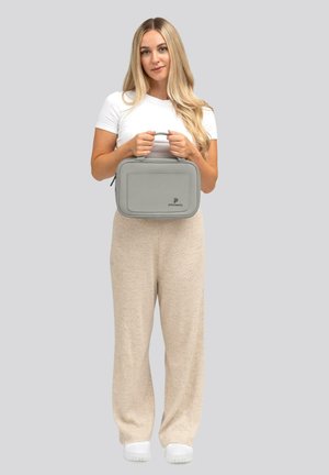 Sac rectangulaire gris avec une poignée supérieure, en matériau lisse, dotée d'une poche frontale et d'un logo de marque. Le modèle porte un pantalon beige texturé et des chaussures blanches.