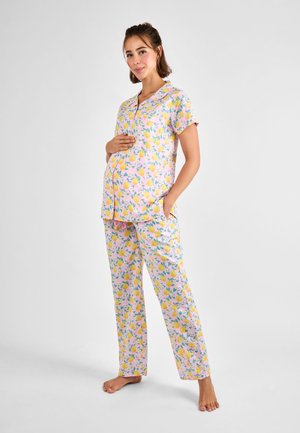 Zwangere vrouw staat blootsvoets, draagt een roze kortemouws bloesje met knopen en een broek met gele bloemenprint, hand op de buik, glimlachend.