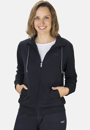 Lächelnde Frau mit kurzen Haaren, die einen schwarzen Hoodie mit Reißverschluss und passende Hose trägt, Hände in den Taschen, darunter ein weißes Hemd.