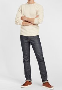 Krämbs strumpor, mörka denimjeans och bruna sneakers. Tröjan har en ribbad halsringning och mudd, jeansen har en rak passform med synliga sömmar.