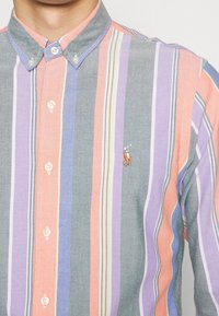 Randig skjorta med knäppning i pastellfärger (rosa, lila, grå, gul) med krage och broderad logotyp på bröstet.