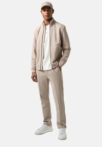 Beige ritsjack met hoge kraag, bijpassende tapered broek en witte sneakers. Glad materiaal met nauwkeurige stikdetails.