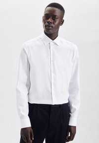 Camicia bianca a maniche lunghe con colletto classico e polsini abbottonati, realizzata in tessuto liscio, abbinata a pantaloni scuri.