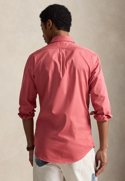 Άνδρας που φοράει κοραλί μακρυμάνικο πουκάμισο με τα μανίκια γυρισμένα και λευκό παντελόνι, γυρισμένος με την πλάτη σε ουδέτερο φόντο.
