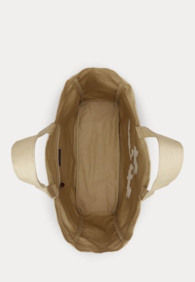 Μπεζ καμβά τσάντα ώμου με φαρδιές λαβές, το ανοικτό πάνω μέρος δείχνει μια εσωτερική τσέπη και λευκή κεντητή ανθοδέσμη στο εσωτερικό μέρος.