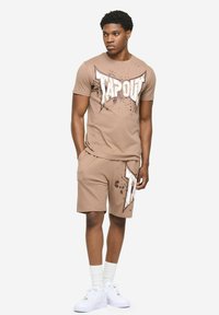 Bež komplet majice kratkih rukava i kratkih hlača s istaknutim natpisom "TAPOUT" u bijeloj boji, ukrašen detaljima prskanja. Komplet nadopunjuju bijele tenisice i čarape.