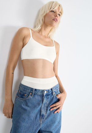 Witte, zachte stof bralette met verstelbare bandjes, gecombineerd met high-waisted, lichtblauwe denim jeans met zichtbare stiksels en knoopsluiting.