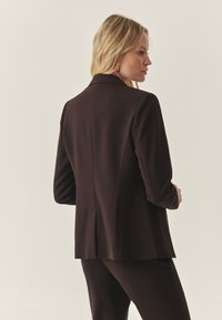 Brun skræddersyet blazer med slået krave, strukturerede skuldre og en enkelt slidse bagpå. Glat stof med en tætsiddende pasform.
