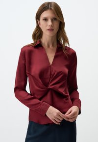 Burgundy satin blouse with a collared neckline and a twisted knot detail at the front. Long sleeves and a slightly curved hem.