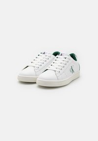 Hvite lave sneakers laget av lær med grønne detaljer, har snørebånd, en jevn tekstur og CK-merke på siden.