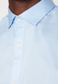 Camisa abotonada de color azul claro con cuello, con botones blancos y una textura suave de algodón. Detalles de costura visibles cerca de los botones.