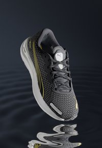 Baskets de course noires avec un motif géométrique, accents jaunes, empeigne en maille et semelle épaisse blanche, comprenant des lacets et un logo sur la languette.